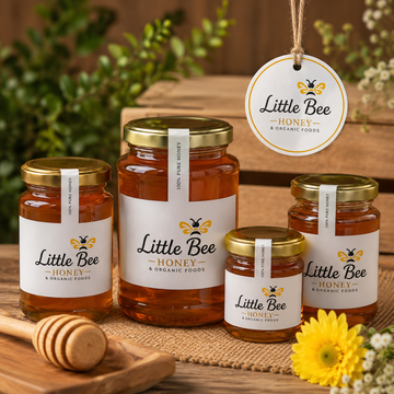 Four glass jars of Little Bee Organic Foods honey in various sizes, arranged on a burlap surface with a wooden honey dipper, greenery in the background, and a hanging brand tag.